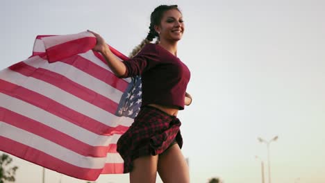 Junges-Glückliches-Amerikanisches-Mädchen,-Das-Läuft,-Während-Es-Die-Amerikanische-Flagge-Hält-Und-In-Die-Kamera-Schaut-1
