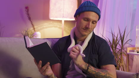 Man-Sitting-On-Sofa-And-Holding-A-Snake-Around-His-Neck-While-Reading-A-Book-At-Home