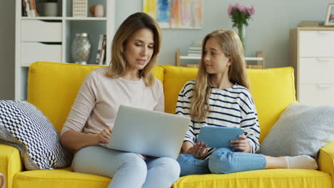 Madre-Rubia-E-Hija-Sentadas-En-Un-Sofá-Amarillo-Mientras-Hablan-Y-Usan-Una-Computadora-Portátil-Y-Una-Tableta-En-La-Sala-De-Estar-1