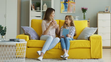 Blonde-Woman-Holding-A-Cup-Of-Coffee-And-Talking-With-Her-Teen-Daughter-Who-Is-Holding-A-Tablet