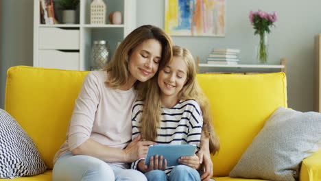 Madre-Rubia-E-Hija-Descansando-En-El-Sofá-De-Casa-Y-Viendo-Algo-En-La-Tableta,-Luego-La-Madre-Abraza-A-Su-Hija