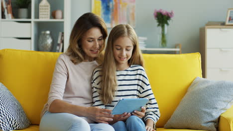 Blonde-Mother-And-Daughter-Making-A-Video-Call-With-A-Tablet-While-Sitting-On-Sofa-In-Living-Room