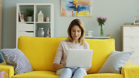 Blonde-Woman-Working-On-The-Laptop-Sitting-On-Sofa-In-Living-Room-1