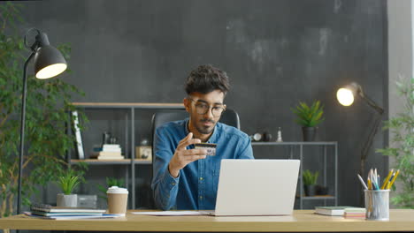 Joven-árabe-Con-Anteojos-Sentado-En-El-Escritorio,-Con-Tarjeta-De-Crédito-Y-Comprando-En-Línea-En-Una-Computadora-Portátil