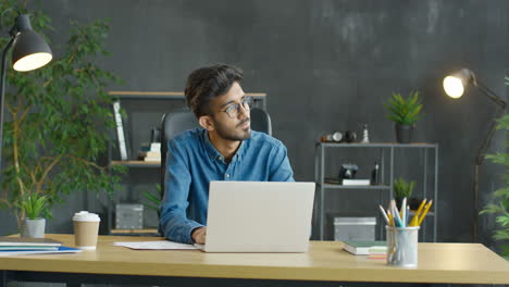 Young-Arabic-Man-Working-Sitting-At-Desk-With-Coffee-To-Go-And-Stationery-In-Front-Of-Laptop-In-The-Office