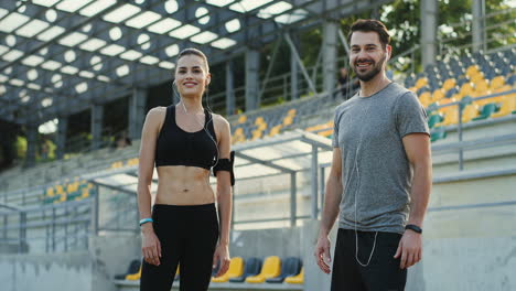 Junge-Joggerpaare-Schauen-Sich-An-Und-Lächeln-Dann-In-Die-Kamera,-Während-Sie-Am-Sommertag-Im-Stadion-Trainieren