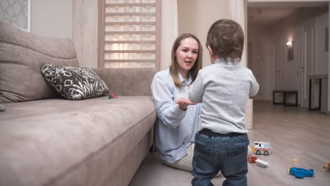 Baby-Unternimmt-Seine-Ersten-Schritte-In-Der-Nähe-Des-Sofas,-Um-Ein-Feuerwehrautospielzeug-Zu-Nehmen-Und-Es-Seiner-Mutter-Zu-Geben