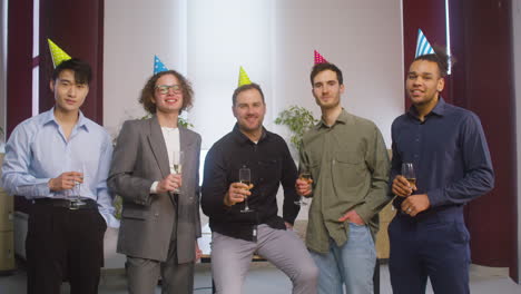 Glückliche-Multiethnische-Gruppe-Männlicher-Kollegen,-Die-Champagnerglas-Halten,-Posieren-Und-Auf-Der-Büroparty-1-In-Die-Kamera-Schauen