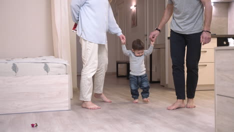 Parents-Holding-Their-Baby's-Hands,-Helping-Him-Take-His-First-Steps-At-Home-1