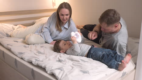 Eltern-Spielen-Mit-Seinem-Sohn-Auf-Dem-Bett,-Während-Das-Baby-Ein-Spielzeugherz-Hält