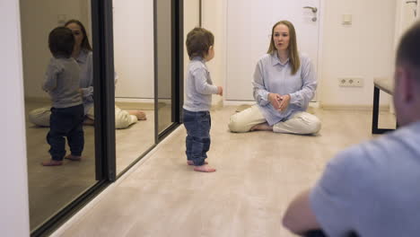 Madre-Y-Su-Bebé-En-Un-Extremo-Del-Pasillo-En-Casa-Mientras-El-Padre-Anima-Al-Bebé-A-Caminar-Hacia-El-Otro-Extremo-1