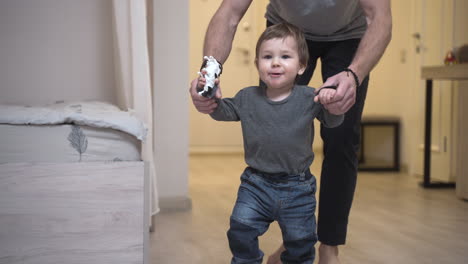 Father-Helping-Her-Baby-To-Take-His-First-Steps-From-The-Corridor-To-The-Living-Room-At-Home-1