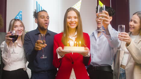 Portrait-Of-A-Woman-Looking-At-The-Camera,-Holding-A-Birthday-Cake-And-Blowing-Out-Candles-Surronded-By-Multiethnic-Colleagues-Who-Raising-A-Toast-At-The-Office-Party