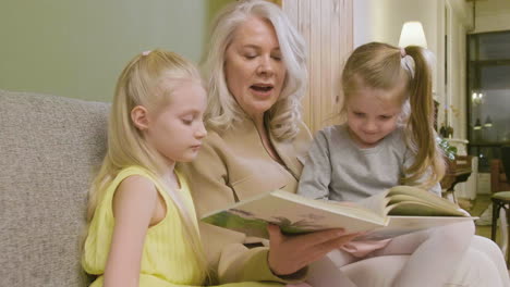 Grandmother-Sitting-On-Sofa-With-Her-Two-Granddaughters-And-Reading-Them-Fairy-Tales-1