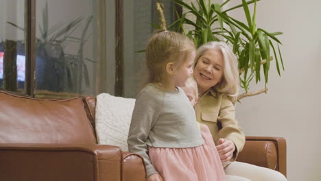 Glückliche-Großmutter-Sitzt-Auf-Dem-Sofa-Und-Umarmt-Ihre-Beiden-Kleinen-Enkelinnen-Zu-Hause