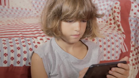 An-attentive-little-girl-browsing-her-smartphone-while-sitting-near-the-bed