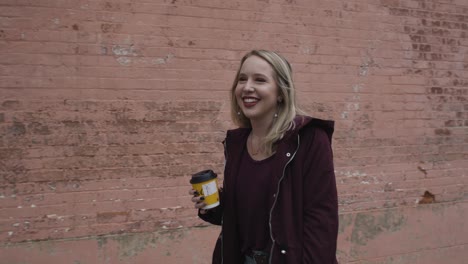 Slow-Motion-Shot-of-a-Carefree-Young-Female-Smiling-and-Walking-Down-the-Street-with-a-Hot-Beverage-in-Hand-and-a-Red-Brick-Wall-in-the-Background