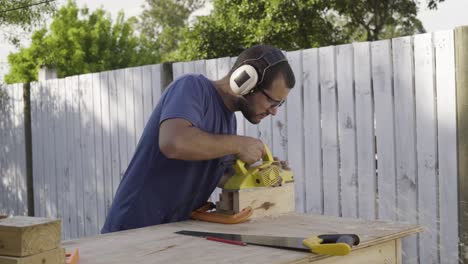 Carpenter-working-outdoors-with-power-tool-electric-planer-cutting-a-block-of-wood