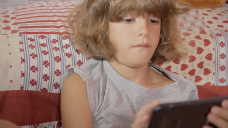 A-cute-little-girl-browsing-her-smartphone-while-sitting-near-the-bed,-looking-tired