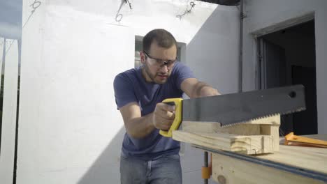 Young-carpenter-craftsman-wears-eye-protection-while-sawing-through-a-piece-of-wood-at-his-workshop