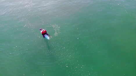 Einsamer-Surfer-Im-Roten-Neoprenanzug-Rashguard-Wartet-Auf-Eine-Welle,-Während-Eine-4k-drohne-Aus-Der-Luft-Dröhnt---Kippt-Bis-Zum-Sonnenaufgang-über-Dem-Pazifischen-Ozean-Am-Jagdton-Beach,-Kalifornischer-Surfwettbewerb