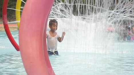 A-mixed-raced-young-boy-exploring-a-children's-water-park-and-running-through-water-spray-as-he-wipes-his-face