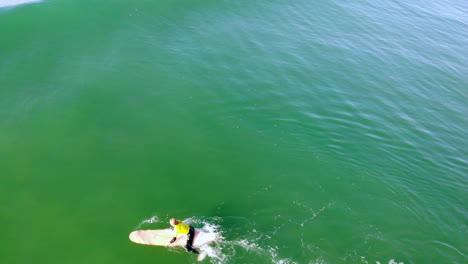 Surfer-Zander-Adelsohn-paddles-out-to-catch-a-wave-in-yellow-wetsuit-at-Huntington-Beach-pier,-California-surfing-competition-in-Pacific-Ocean-with-4k-aerial-DJI-drone-fly-over---tilt-down