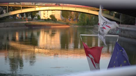 Cámara-Lenta-De-Banderas-En-La-Parte-Superior-Del-Barco-Mientras-Navega-En-El-Canal-Del-Danubio-En-Viena,-Austria