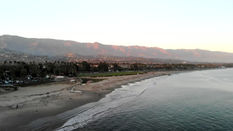 Aerial-drone-shot-at-sunset-above-a-Pacific-Ocean-beach-and-marina-looking-over-the-the-beautiful-city-of-Santa-Barbara-and-the-hills-of-Monecito,-California