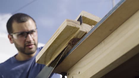 Low-angle-shot-of-tradesman-wearing-safety-glasses-and-cutting-wood-with-a-saw