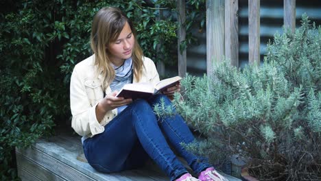 Attraktives-Teenager-Mädchen-Liest-Buch-Auf-Der-Terrasse-Zwischen-Pflanzen