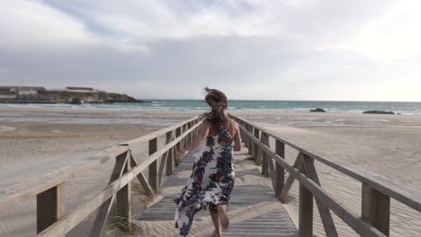 Young-woman-runs-away-from-camera,-running-down-wooden-boardwalk-on-sandy-beach-in-Gibraltar,-Spain