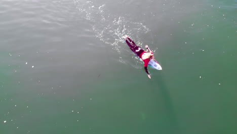 Male-surfer-Zander-Adelsohn-in-red-wetsuit-paddles-in-Pacific-Ocean-at-Huntington-Beach,-California-taken-fro-DJI-4k-aerial-drone