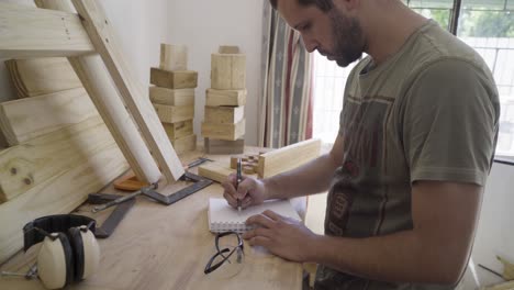 Young-male-carpenter-plans-out-project-drawing-in-a-sketchbook-at-his-workbench-work-desk