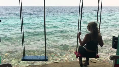 Young-beautiful-caucasian-woman-enjoying-the-view-on-a-swing-in-front-of-the-ocean-crystal-clear-blue-water