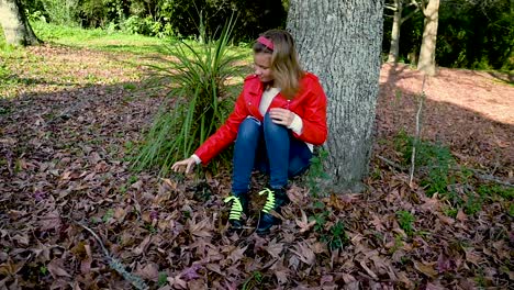 Attraktives-Teenager-Mädchen,-Das-Unter-Einem-Baum-Sitzt-Und-Mit-Herbstlaub-Spielt