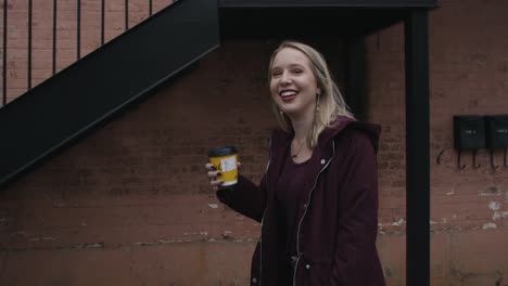 Slow-Motion-of-a-Young-Woman-Smiling-and-Walking-as-She-Raises-Her-Coffee-Cup-Towards-the-Camera-with-a-Red-Brick-Wall-and-Staircase-in-the-Background