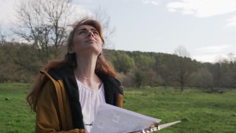 Young-woman-with-notes-in-hand-walks-on-path-on-nature-trail-and-looks-at-the-sky-to-see-if-there-is-something-to-discover