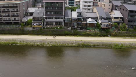 Aerial-view-above-female-cycling-on-a-bike-along-river-in-Kyoto-city-buildings-district