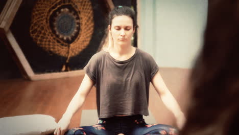 Aged-1970s-film-effect,-slow-motion-handheld-shot:-a-young-woman,-during-a-yoga-lesson,-sitting-on-the-floor,-repeating-a-position-following-the-teacher's-voice,-eyes-closed