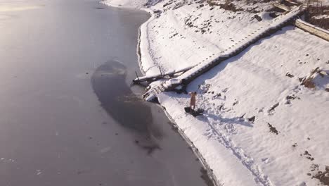 Drone-descending-near-shore-of-frozen-lake-with-man-drying-off-using-towel-after-ice-swimming