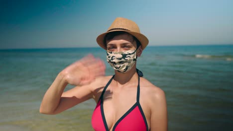 Young-bikini-woman-on-the-beach-wearing-face-mask-and-sunhat-waving