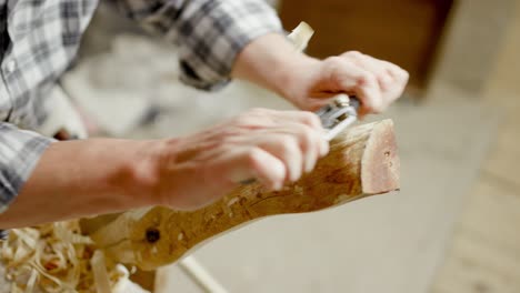 Man-in-checkered-shirt-uses-handplane-to-slice-thin-layer-of-wood-off-of-wooden-workpiece,-slow-motion,-close-up