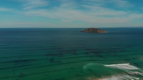 Olas-Ondulantes-Con-Una-Pequeña-Isla-Remota-En-El-Fondo,-Rocas-En-Una-Playa-De-Arena,-Cielos-Azules-Con-Algunas-Nubes