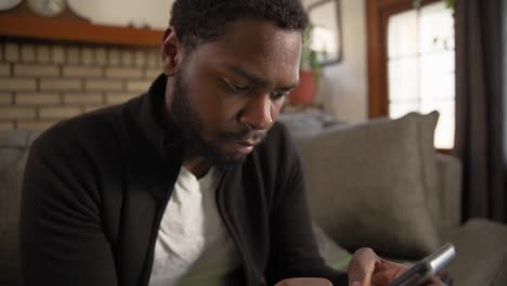 Black-man-texting-on-phone-concentrates-in-living-room-close-up-tilt