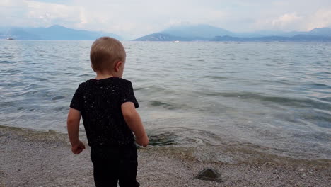 Little-boy-throwing-rocks-in-the-water-in-Lake-Garda,-in-Northern-Italy,-on-a-partially-cloudy-September-day