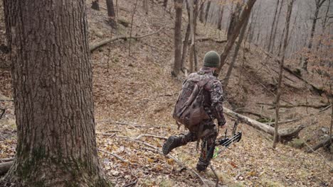 Bowhunter-holding-bow-in-camouflage-outfit-walking-off-trail-in-winter-forest