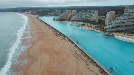 Aerial-dolly-in-of-sea-hitting-sand-shore-near-world-largest-swimming-pool-and-resorts-in-Algarrobo,-Chile