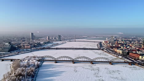 Riga,-Latvia-in-winter-over-the-frozen-Daugava-River-and-bridges---aerial-pull-back-flyover