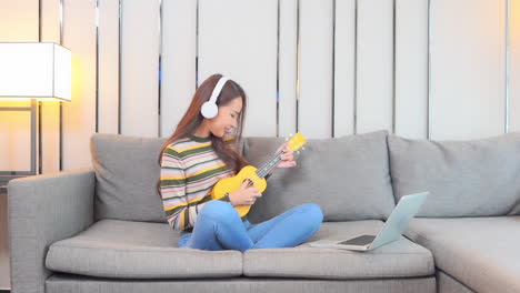 A-cute-young-woman-sitting-cross-legged-on-a-couch-listens-to-music-through-her-headphones-while-practicing-her-ukulele